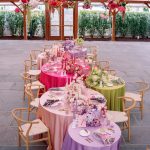 Indoor pavilion reception space with ombre-colored round tables, hanging chandelier adorned with vibrant floral arrangements, and wooden chairs