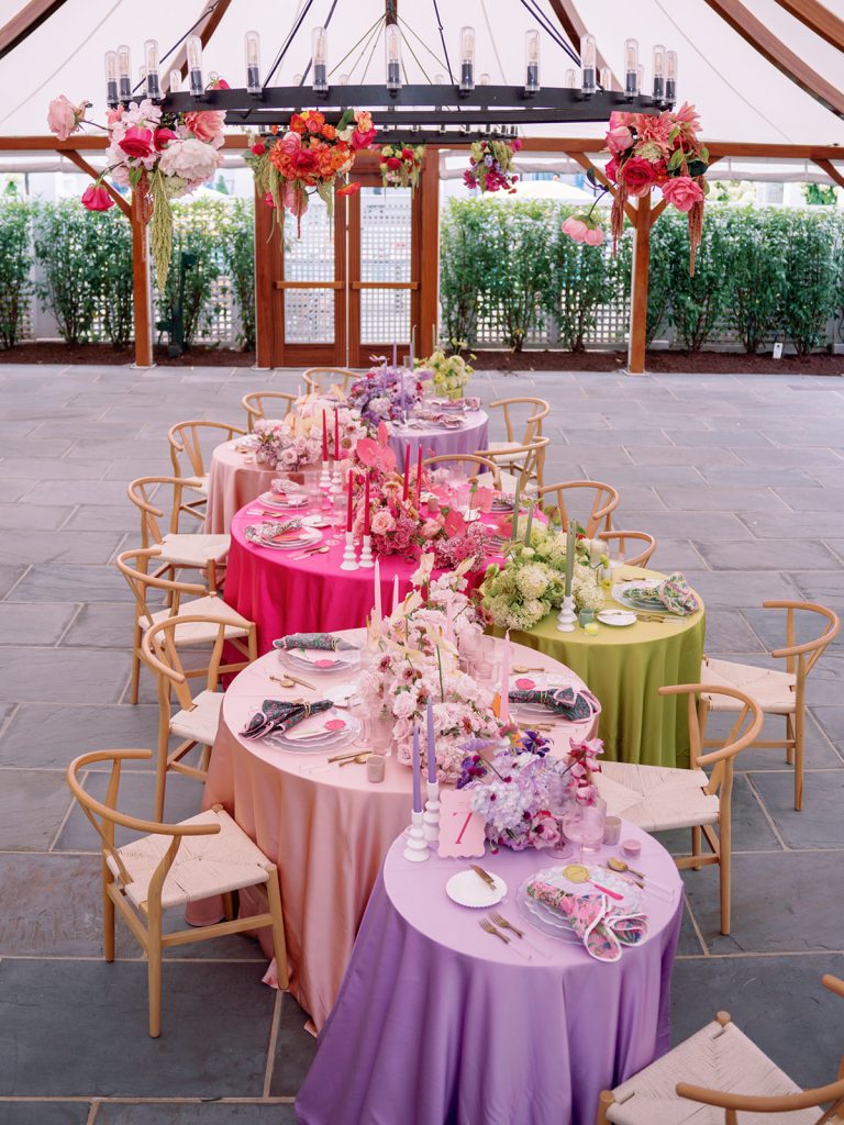 Indoor pavilion reception space with ombre-colored round tables, hanging chandelier adorned with vibrant floral arrangements, and wooden chairs
