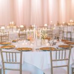 Wedding reception table with gold chargers, tall candles, pink florals, and champagne draping backdrop