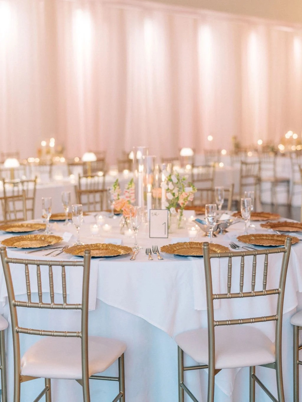 Wedding reception table with gold chargers, tall candles, pink florals, and champagne draping backdrop