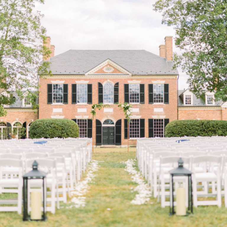 Historic brick mansion with outdoor ceremony setup featuring white folding chairs and lanterns on lawn