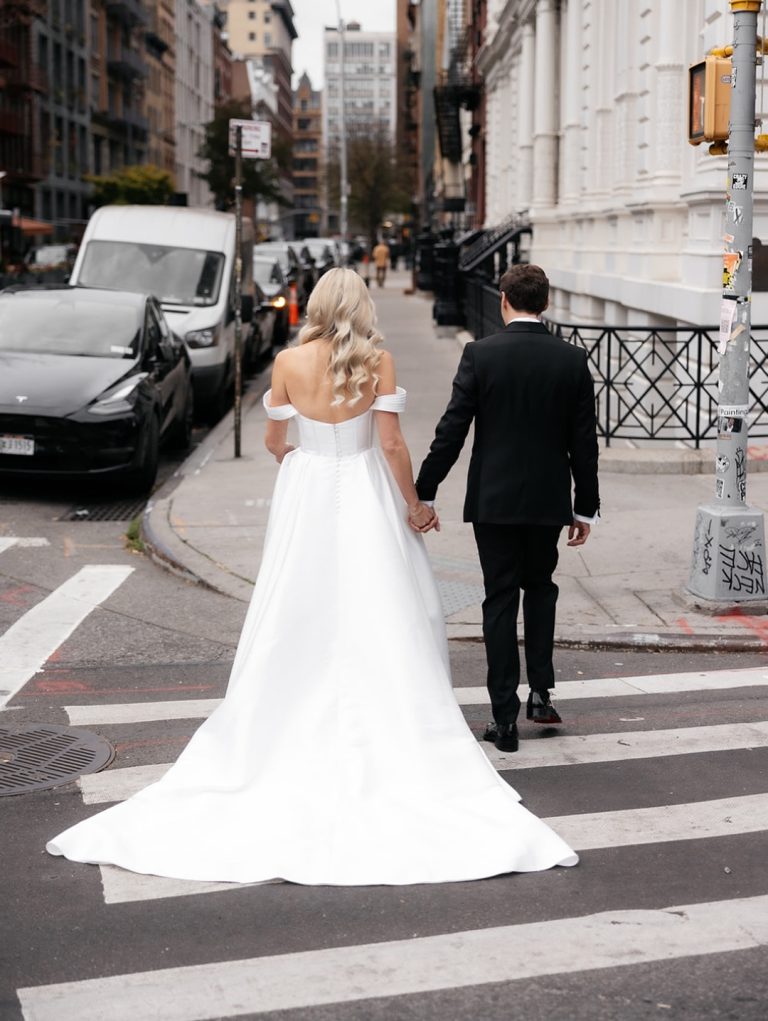 Newlyweds holding hands while crossing urban street, bride's white gown with train flowing behind them