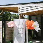 Modern wedding ceremony entrance with custom welcome signage and oversized paper flower decorations