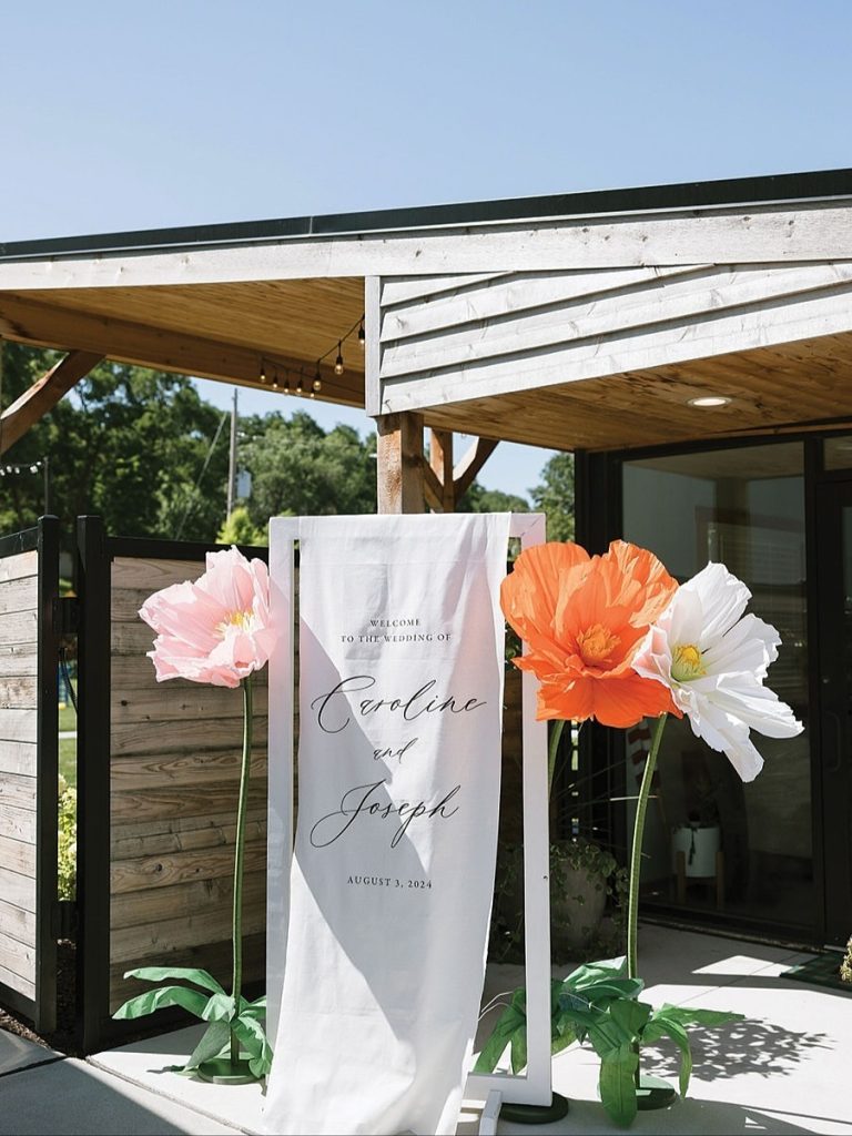 Modern wedding ceremony entrance with custom welcome signage and oversized paper flower decorations