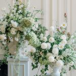 White and green garden-style floral arrangements with dahlias, cosmos, and trailing vines displayed on white pedestals
