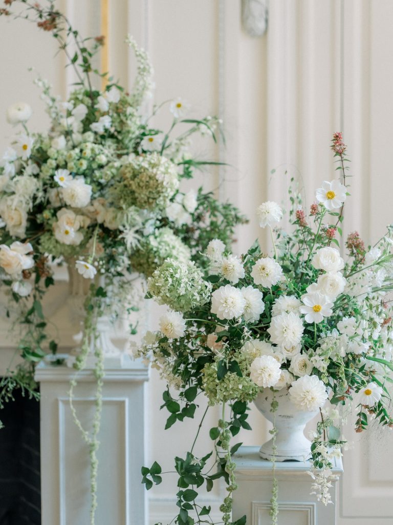 White and green garden-style floral arrangements with dahlias, cosmos, and trailing vines displayed on white pedestals
