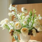 Elegant white and cream floral arrangement with ranunculus, roses, pine cones, and greenery in a white ceramic urn