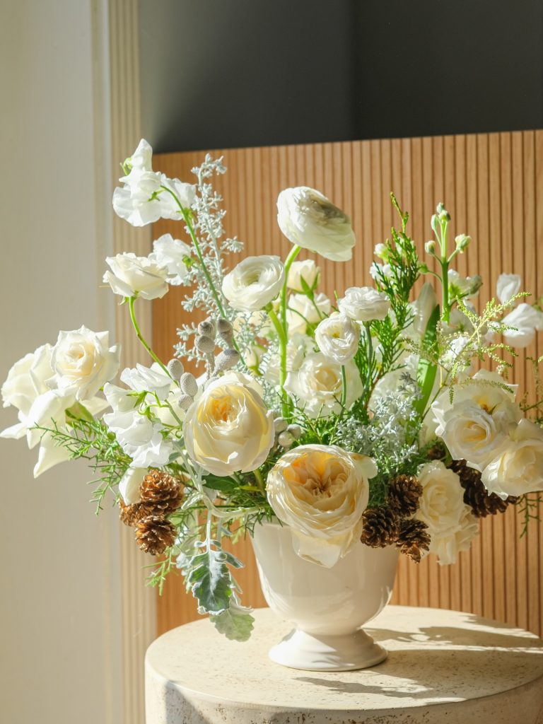 Elegant white and cream floral arrangement with ranunculus, roses, pine cones, and greenery in a white ceramic urn