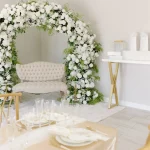 White floral arch behind cream tufted loveseat with modern white and gold event table in minimalist wedding venue