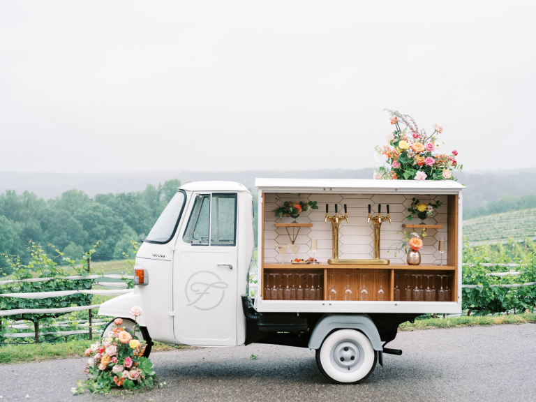White vintage beverage truck converted into mobile bar with floral arrangements and gold tap taps at outdoor wedding venue