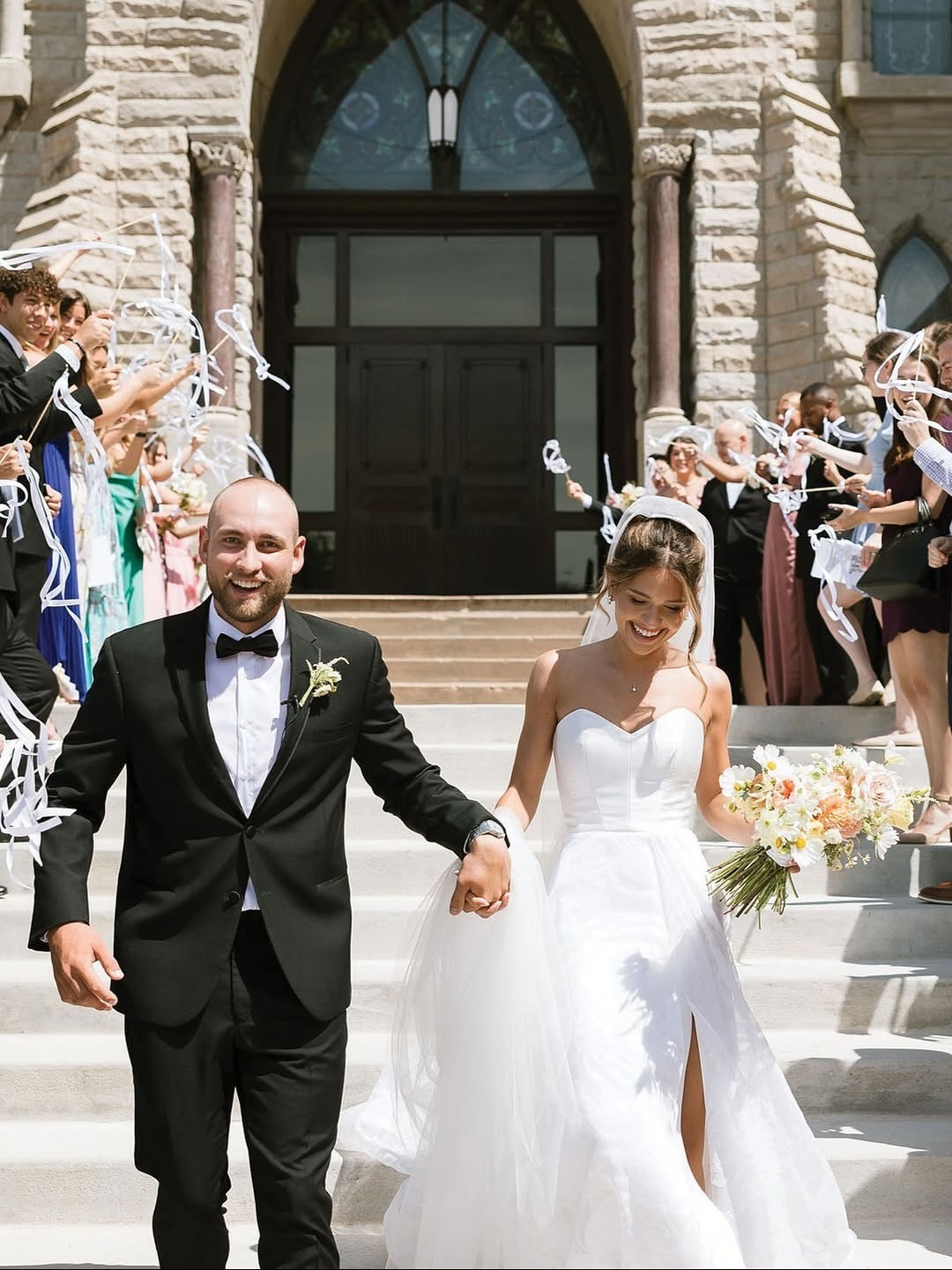 Newlyweds exit historic stone building during ribbon wand send-off with wedding party celebrating around them