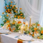 Wedding reception tablescape with orange marigold centerpieces, pillar candles, blue-rimmed china, and mustard velvet runner