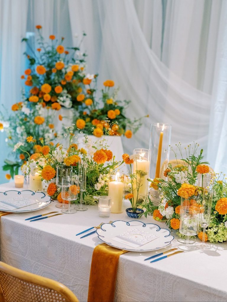 Wedding reception tablescape with orange marigold centerpieces, pillar candles, blue-rimmed china, and mustard velvet runner