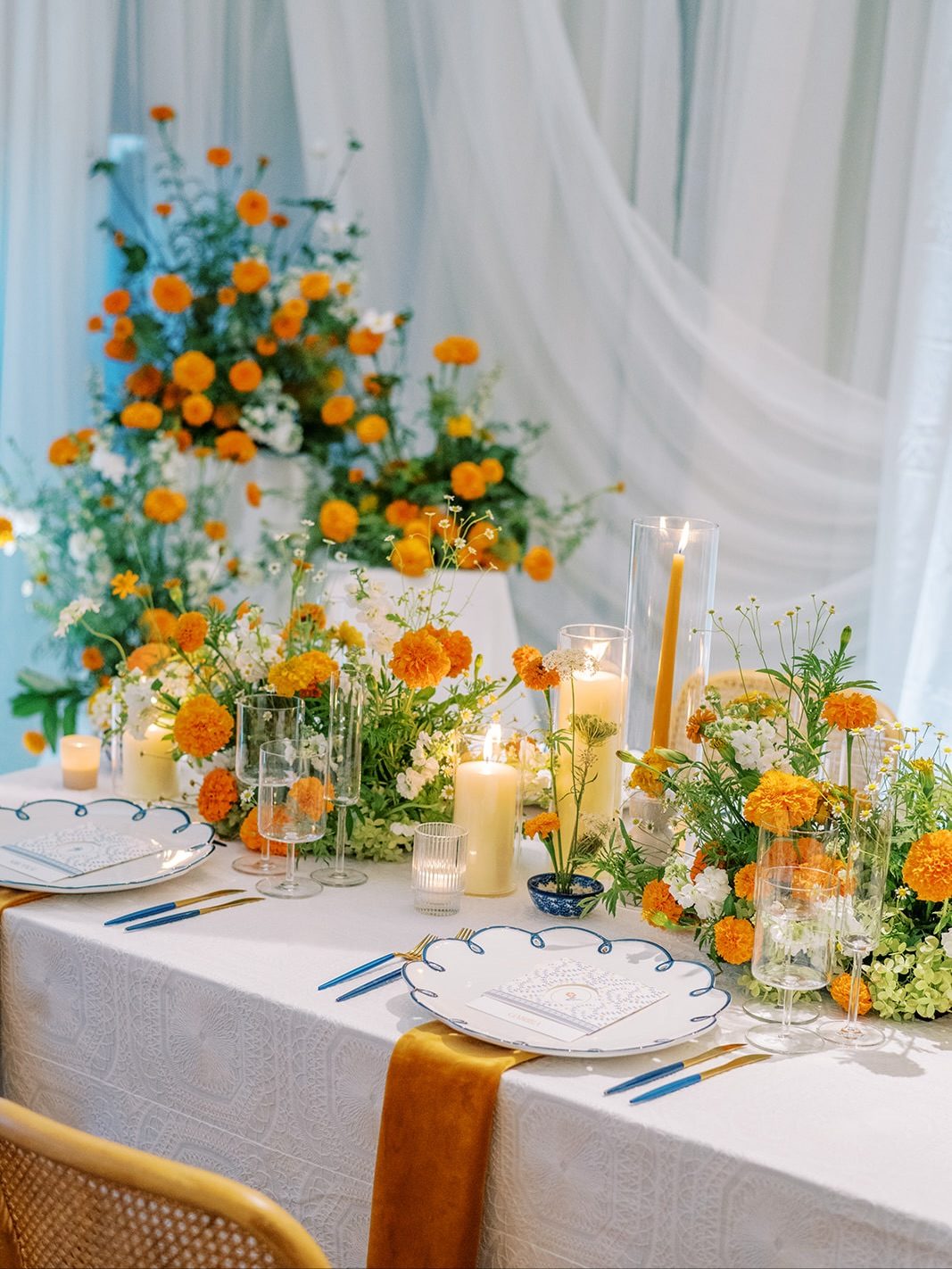 Wedding reception tablescape with orange marigold centerpieces, pillar candles, blue-rimmed china, and mustard velvet runner