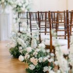 Wedding ceremony aisle lined with brown chiavari chairs and ivory floral arrangements featuring roses, hydrangeas, and greenery