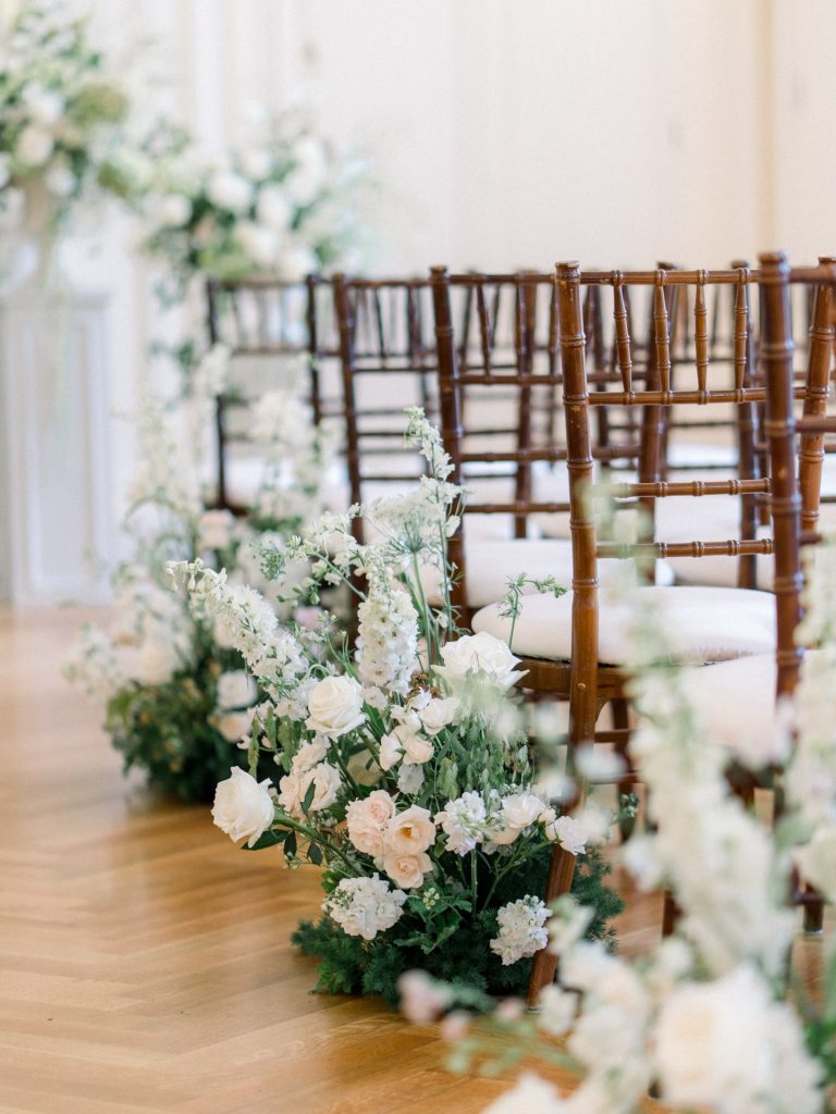 Wedding ceremony aisle lined with brown chiavari chairs and ivory floral arrangements featuring roses, hydrangeas, and greenery