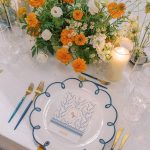 Elegant place setting with blue-rimmed china plate, gold flatware, and orange and cream floral centerpiece with candlelight