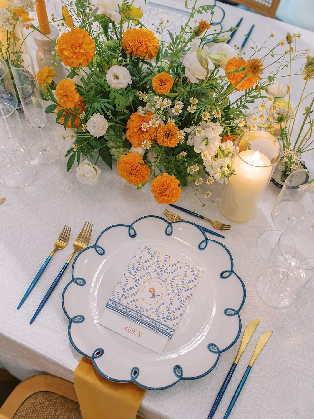 Elegant place setting with blue-rimmed china plate, gold flatware, and orange and cream floral centerpiece with candlelight