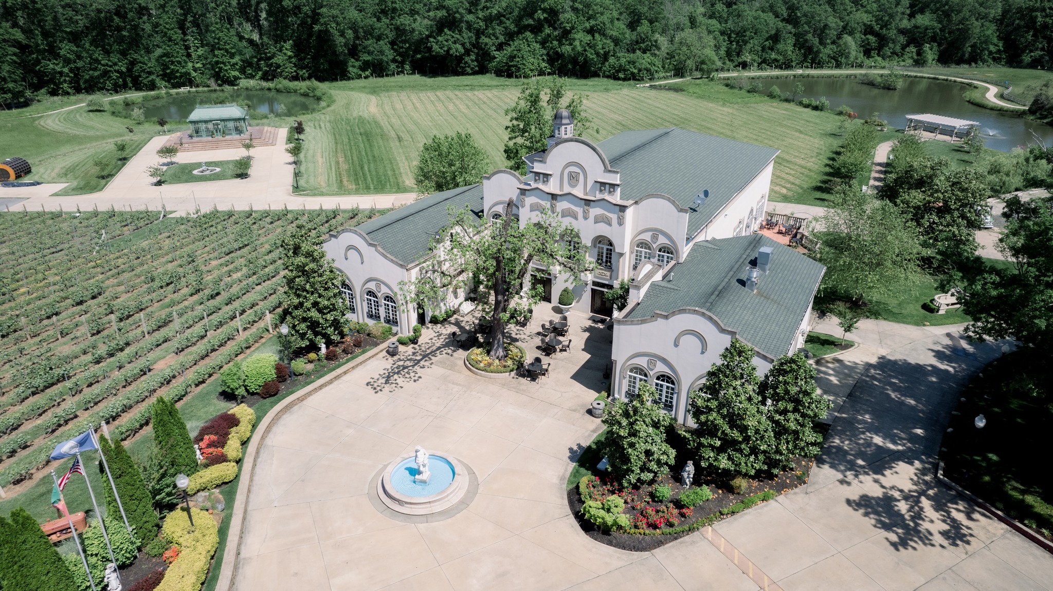 Aerial view of elegant estate wedding venue with white stucco buildings, fountain courtyard, vineyard rows, and pond