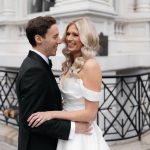 Newlywed couple embracing on city street, bride in off-shoulder gown with groom in black suit