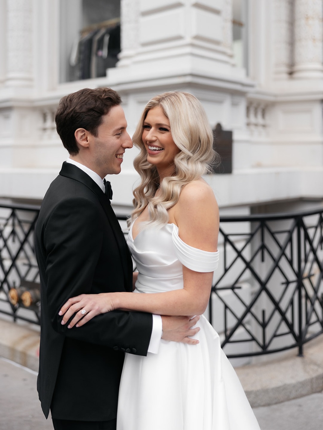 Newlywed couple embracing on city street, bride in off-shoulder gown with groom in black suit