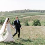 Newlyweds holding hands while walking across open field with rolling vineyard hills in background