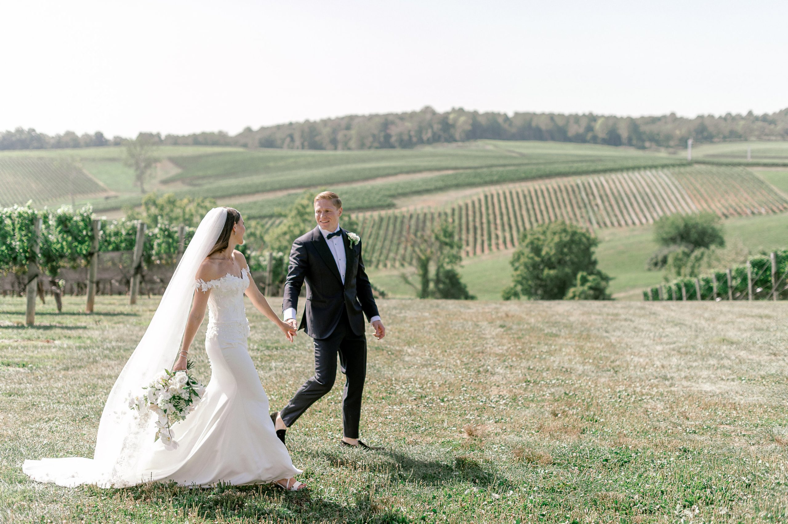 Newlyweds holding hands while walking across open field with rolling vineyard hills in background