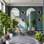 Hotel lobby with teal walls, arched mirrors, copper circular accents, striped floor, and lush tropical plants