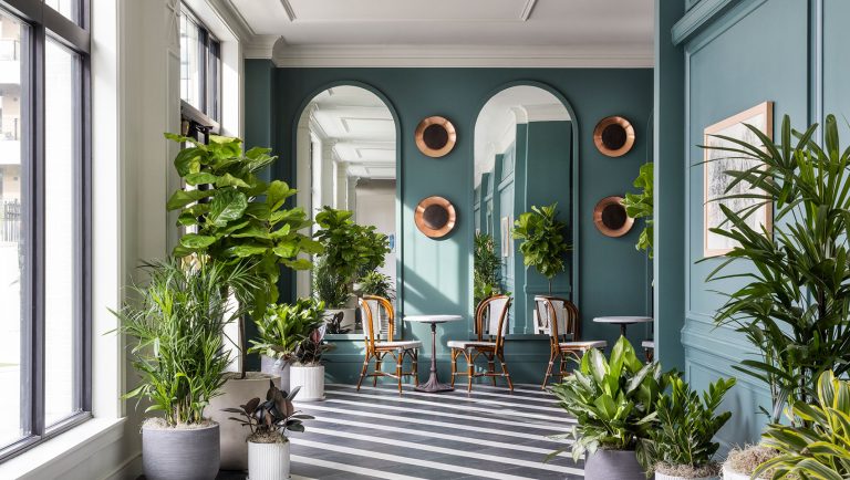 Hotel lobby with teal walls, arched mirrors, copper circular accents, striped floor, and lush tropical plants