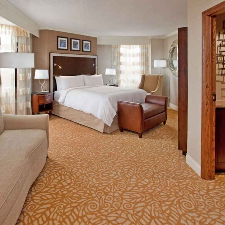 Elegant hotel suite with king bed, leather seating, and gold patterned carpeting