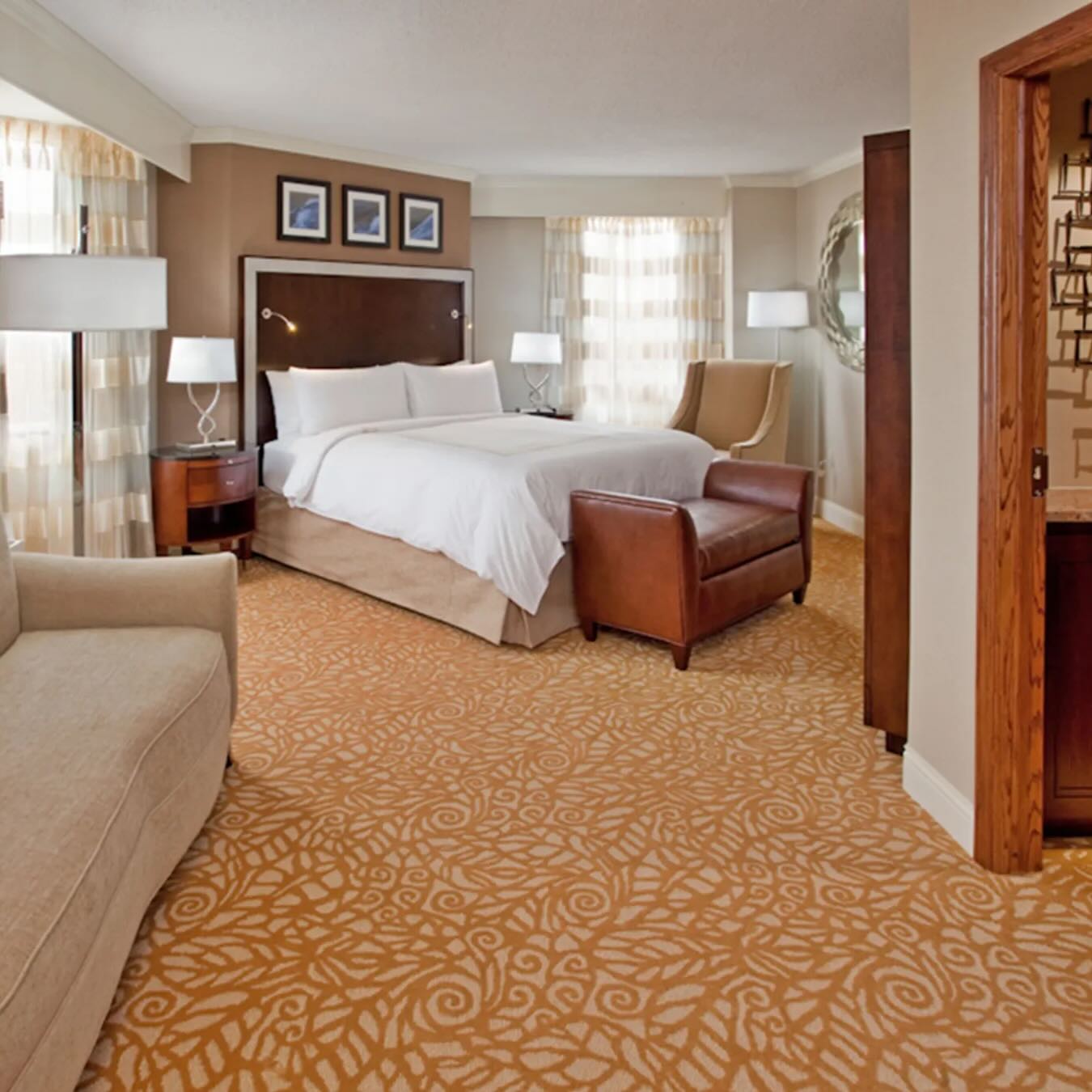 Elegant hotel suite with king bed, leather seating, and gold patterned carpeting