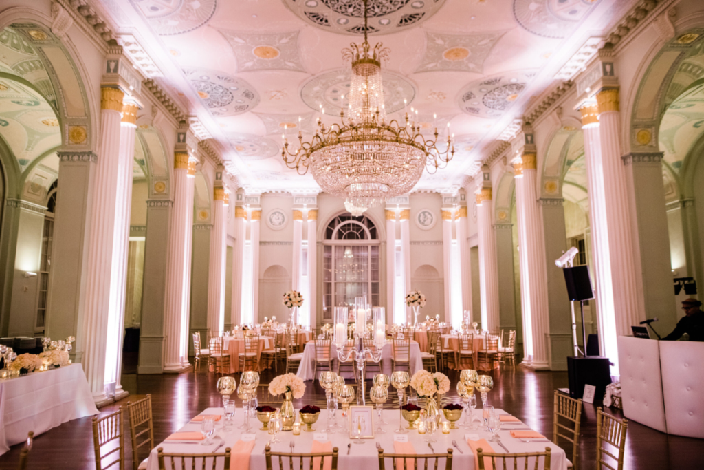 Elegant ballroom wedding reception with ornate crystal chandelier, white columns, and pink uplighting in Alpharetta
