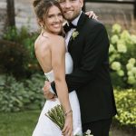 Happy newlyweds posing together outdoors, groom in black tuxedo and bride with loose updo and romantic bouquet