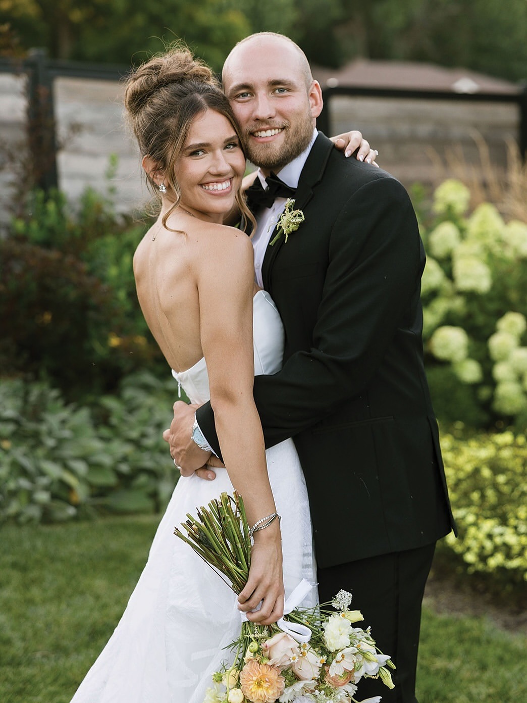 Happy newlyweds posing together outdoors, groom in black tuxedo and bride with loose updo and romantic bouquet