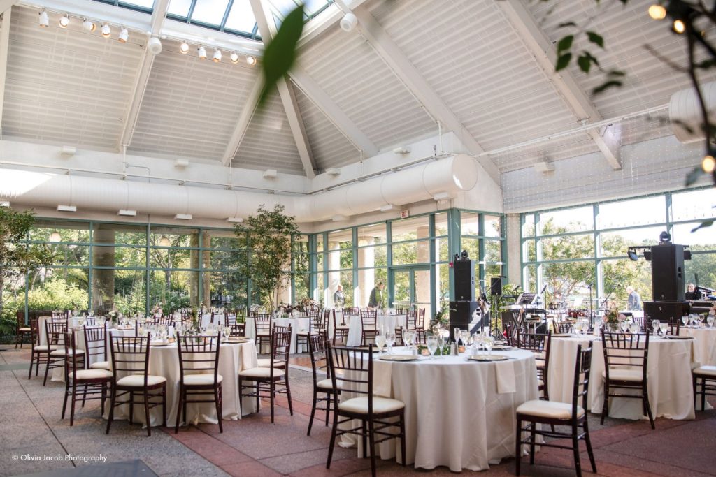 Bright reception hall with vaulted ceilings, floor-to-ceiling windows, mahogany chiavari chairs, and indoor trees creating an elegant Northern Virginia wedding setting