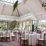 Bright conservatory reception space with vaulted ceiling, floor-to-ceiling windows, and round tables with ivory linens and dark chiavari chairs