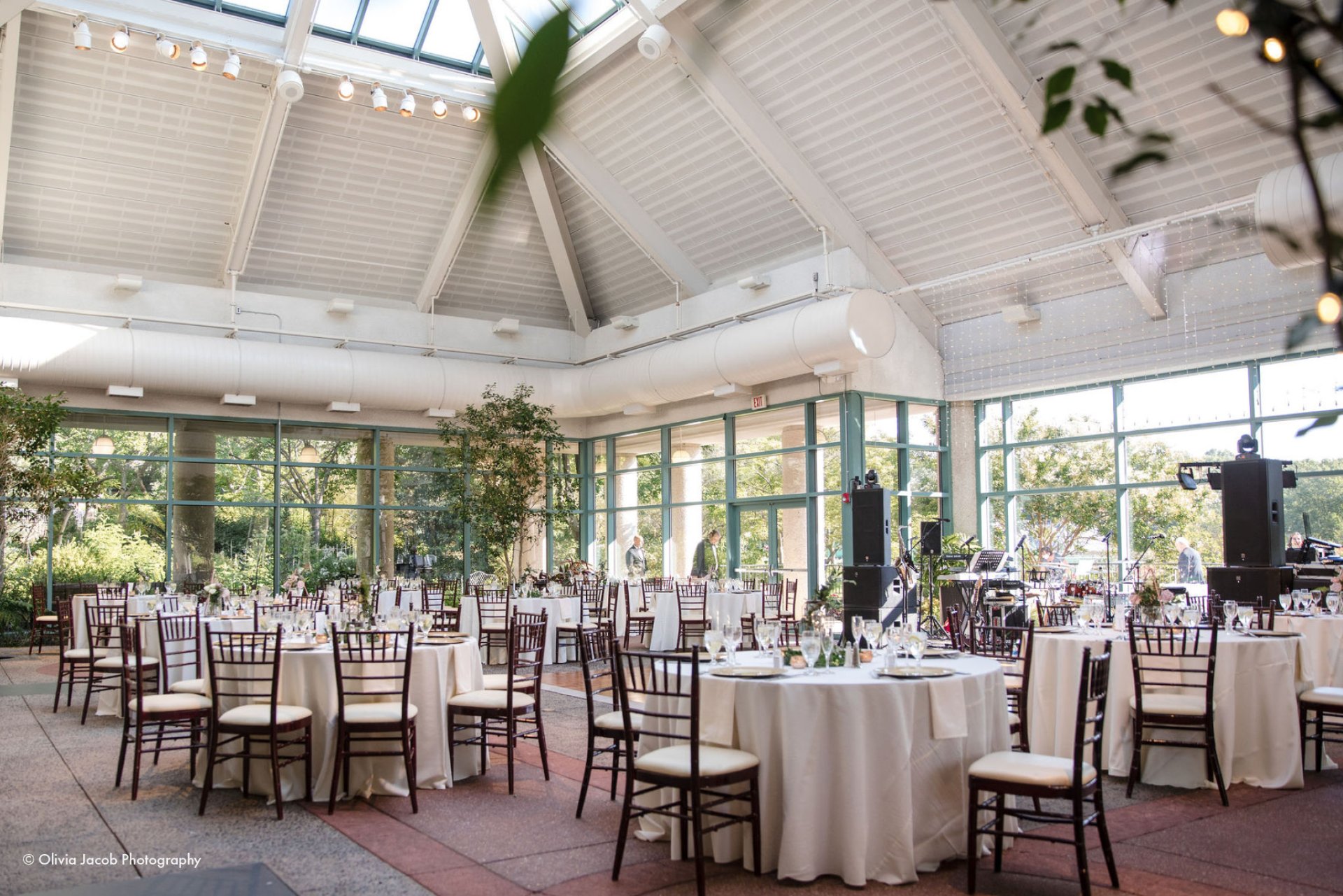 Bright conservatory reception space with vaulted ceiling, floor-to-ceiling windows, and round tables with ivory linens and dark chiavari chairs