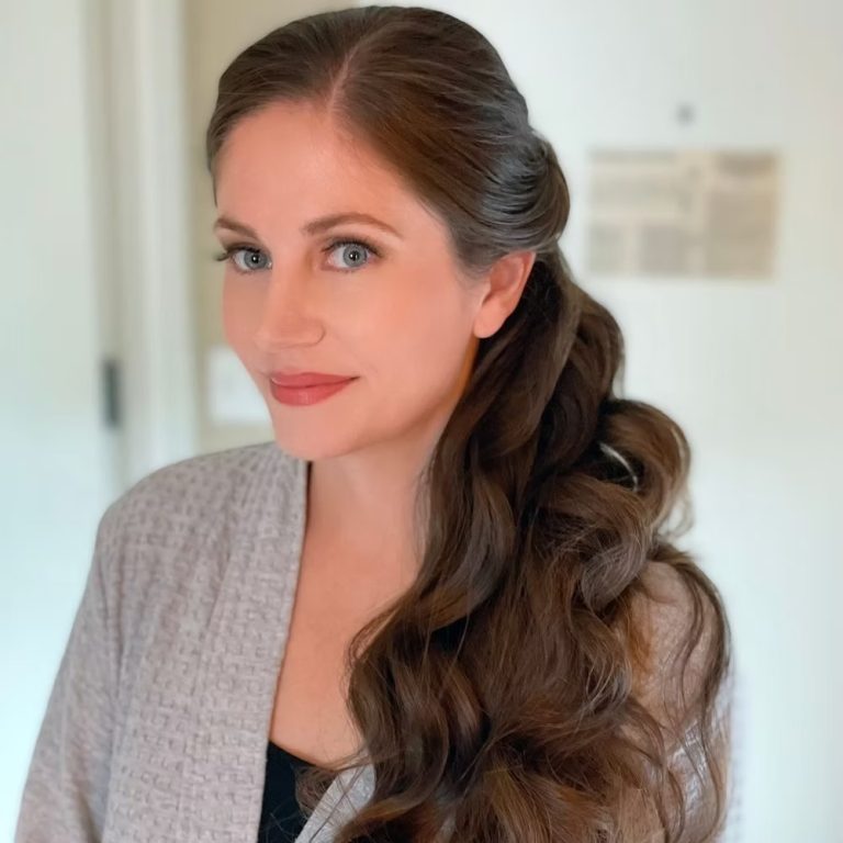 Woman with long wavy brown hair in a side ponytail and gray blazer in a bright interior setting