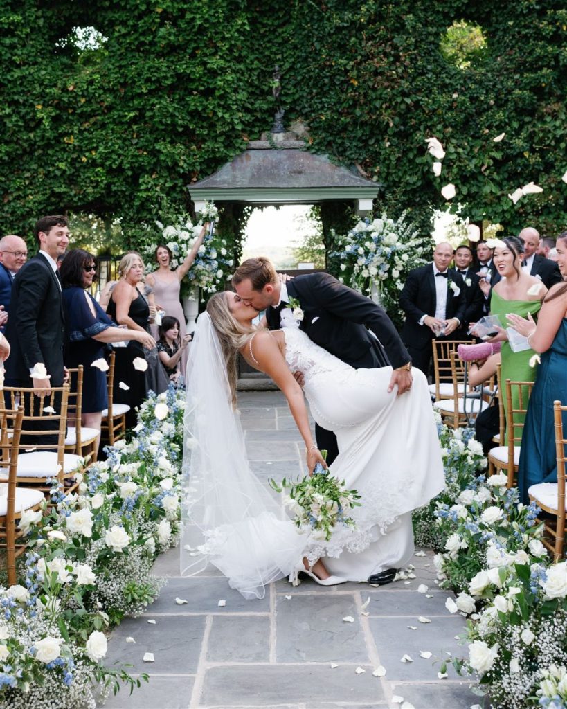 Groom dips bride for kiss during recessional as guests toss petals at ivy-covered gazebo ceremony