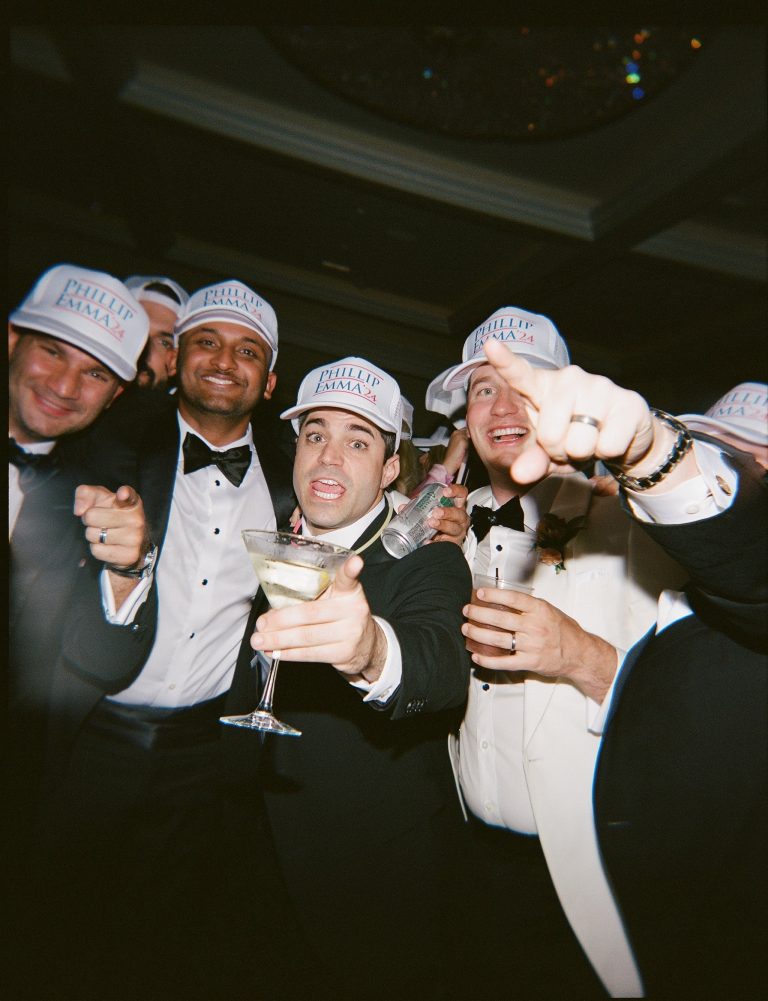 Groomsmen in tuxedos wearing personalized white trucker hats toasting with cocktails at wedding reception
