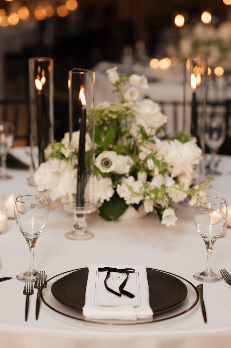 Wedding reception table with white floral centerpiece, tall candles, black place settings, and elegant place cards