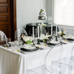 Modern wedding table with clear acrylic ghost chairs, white linens, black plates, and white floral centerpieces