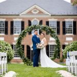 Bride and groom standing under circular greenery arch in front of brick Georgian-style mansion during outdoor ceremony