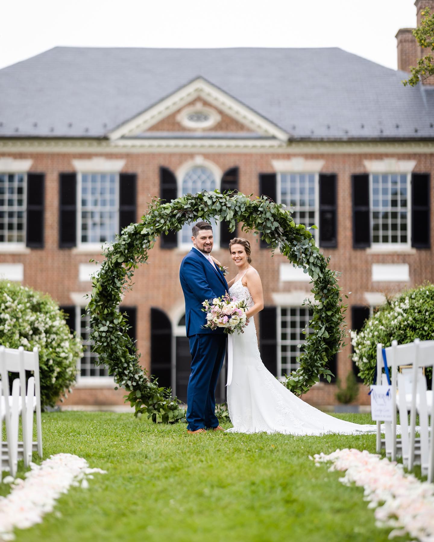 Bride and groom standing under circular greenery arch in front of brick Georgian-style mansion during outdoor ceremony