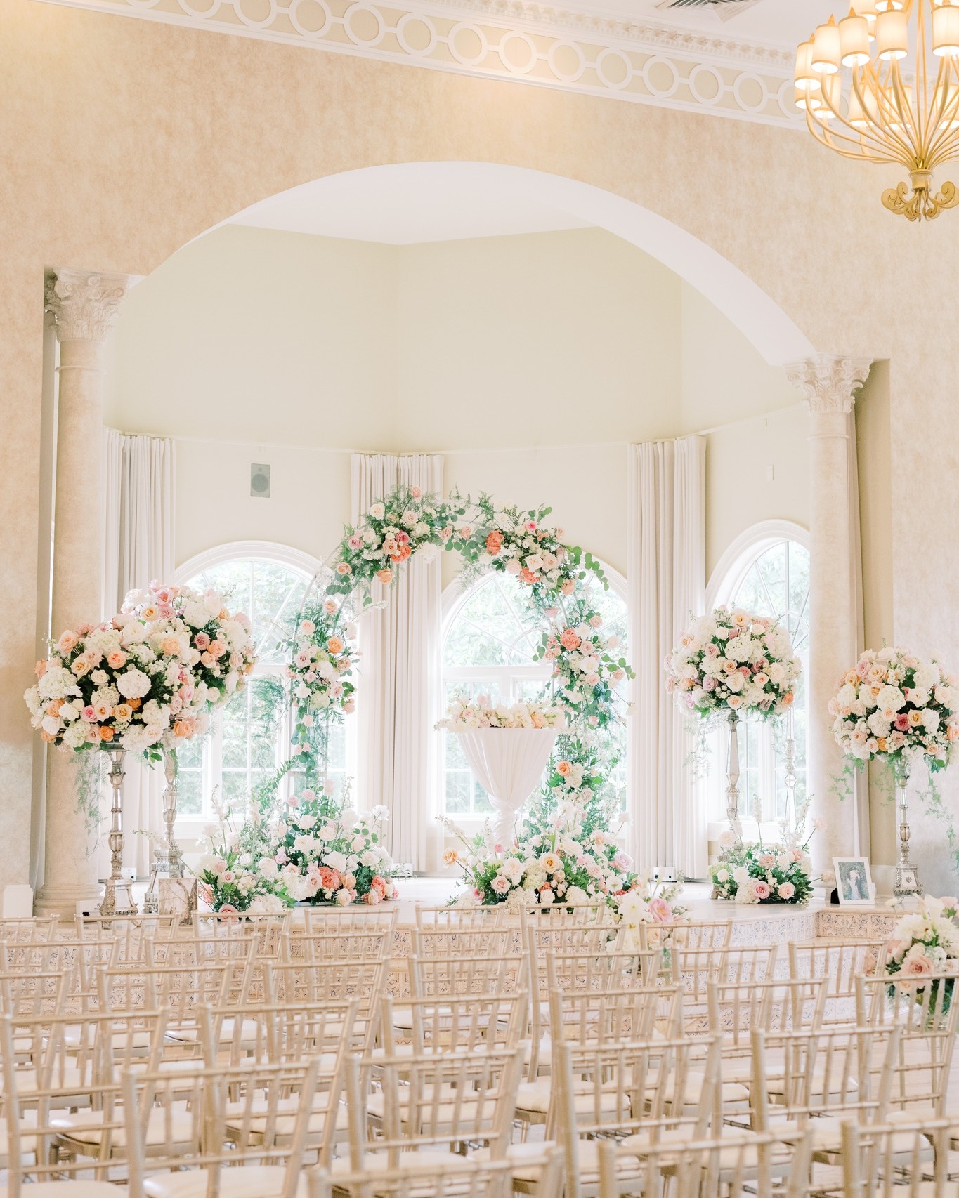 Romantic ballroom ceremony setup with blush floral arch, tall arrangements, and gold chiavari chairs