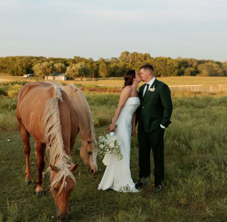 Bride and groom kissing in pasture with grazing horse and tree line at Seven Willows venue