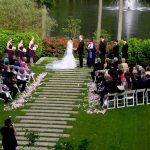 Outdoor wedding ceremony on terraced steps with wedding party, officiant, and seated guests beside pond
