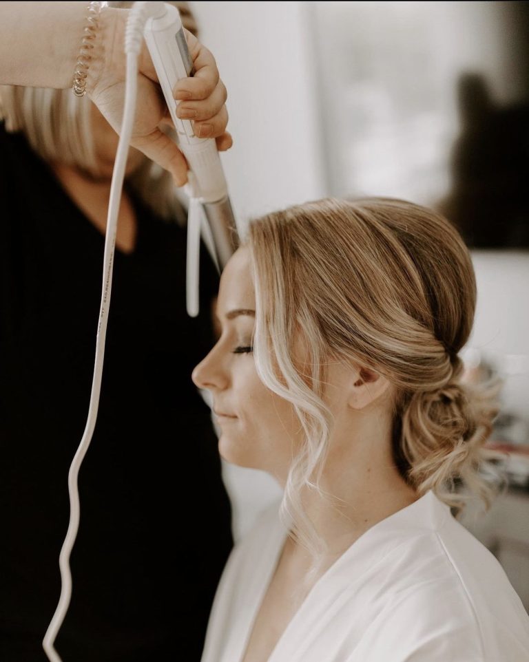 Hairstylist creating romantic updo with loose tendrils for wedding day styling
