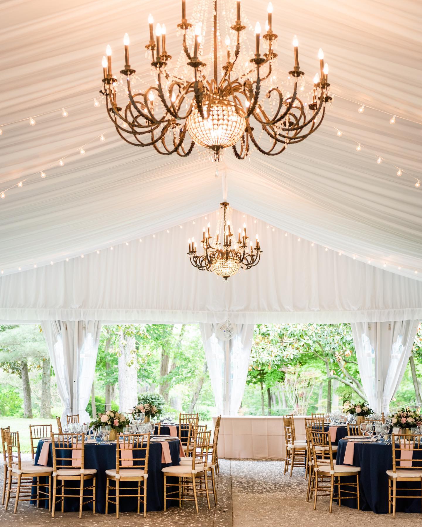 Elegant white draped tent with ornate gold chandeliers and navy and blush reception tables with gold chiavari chairs