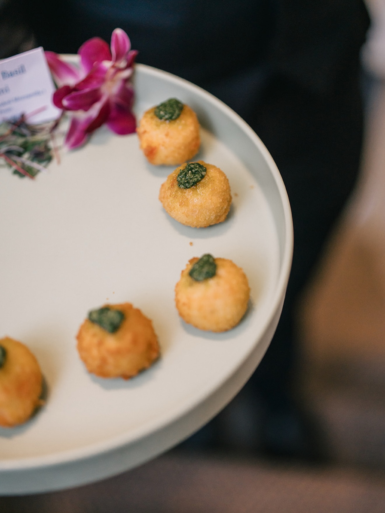 Golden fried appetizer balls topped with green herb sauce on white plate with pink orchid garnish